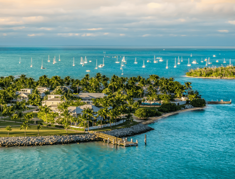 High-angle view of a tropical island in the Florida Keys with luxury waterfront homes, palm trees, and sailboats anchored in turquoise water.