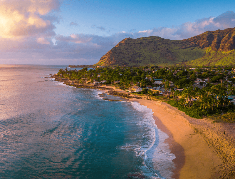 An aerial view of a golden sand beach and turquoise water on the Oahu coast, backed by steep, lush green mountains at sunset.