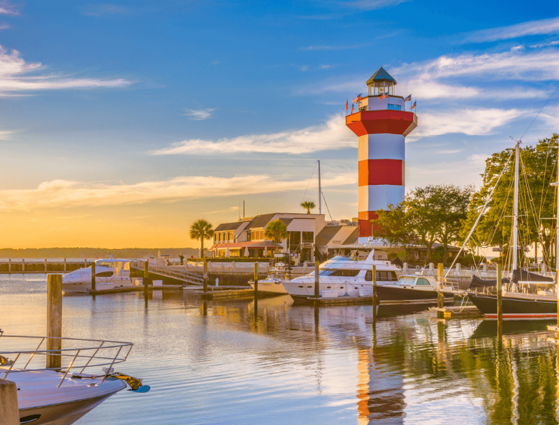 Scenic view of the Harbour Town Lighthouse at sunset in Hilton Head, South Carolina, with yachts and sailboats moored in the marina.