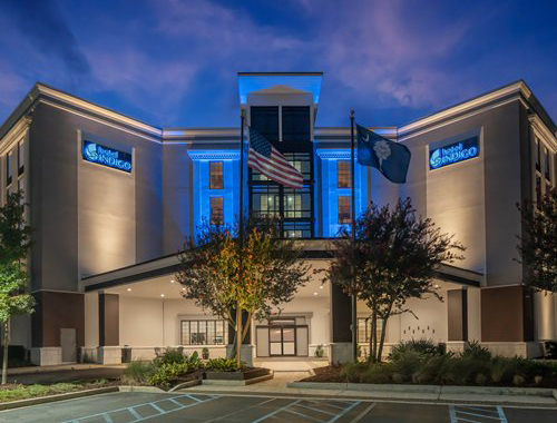 The Hotel Indigo Mount Pleasant exterior at dusk, featuring blue accent lighting on the facade and flagpoles with the American and South Carolina flags.