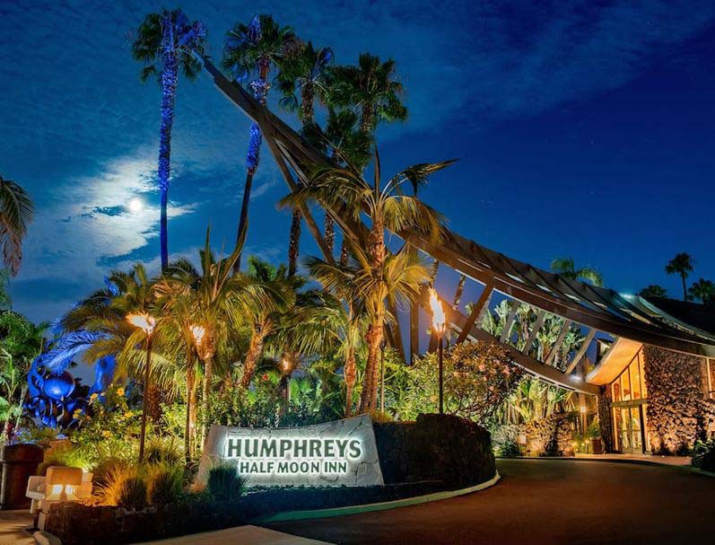 Night view of the entrance to Humphreys Half Moon Inn in San Diego, featuring a lit sign, tiki torches, and palm trees under a moonlit sky.