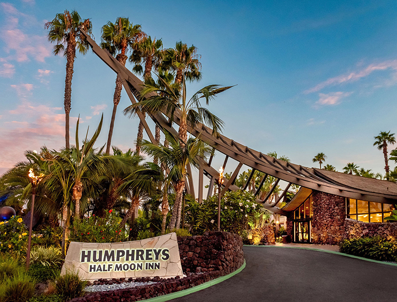 Humphreys Half Moon Inn’s lush resort entrance at dusk, featuring a distinct, curved Polynesian-style roof structure, torch lighting, and prominent signage.