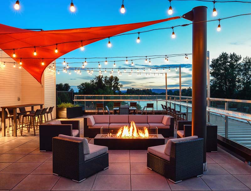 A cozy patio featuring low outdoor seating and a burning fire pit, situated under a bright red canopy and string lighting.