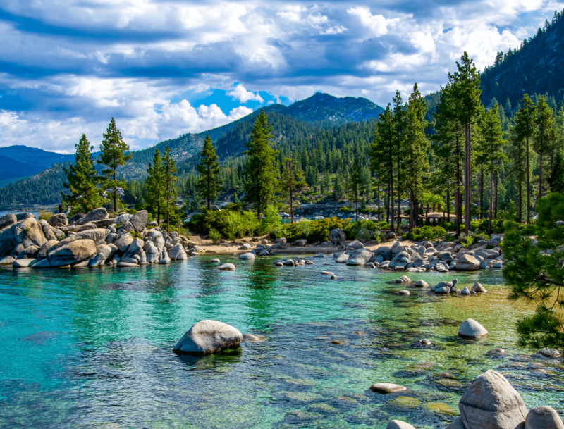 Clear turquoise waters of Lake Tahoe, California, surrounded by large smooth boulders and tall pine trees under a cloudy blue sky.
