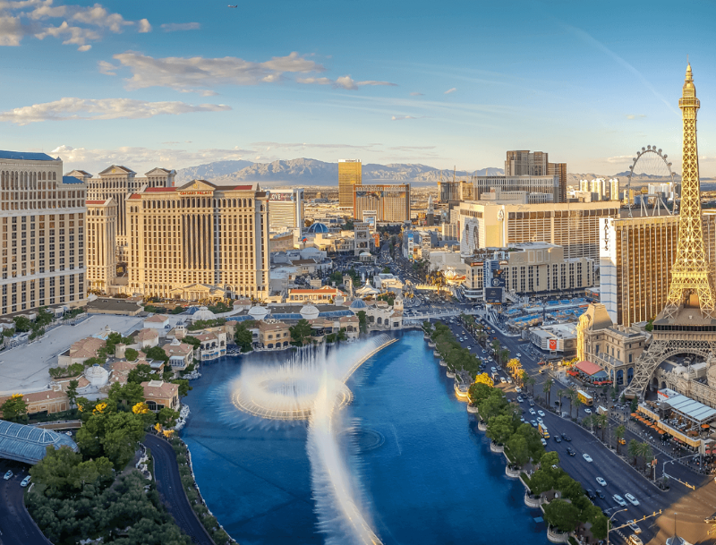 Daytime aerial view of the Las Vegas Strip featuring the Bellagio fountains in mid-show, surrounded by casino resorts and the Eiffel Tower replica.