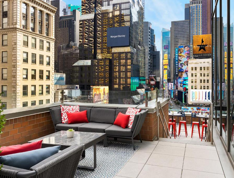 The M Social Hotel rooftop terrace featuring grey sectional seating with red pillows, marble tables, and a glass railing overlooking Times Square.