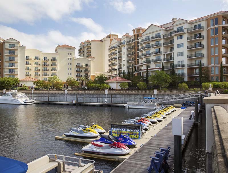 A row of jet skis docked at a marina with lushly-landscaped large resort buildings in the background.