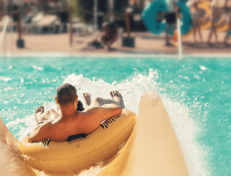 A first-person perspective of a person riding down a yellow water slide on an inflatable tube, splashing into a turquoise pool at a water park.
