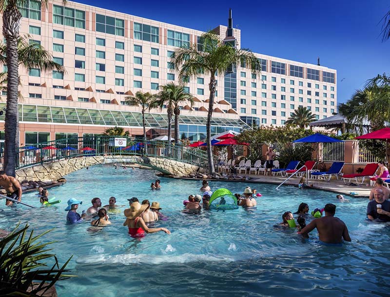 A large swimming pool filled with people enjoying the water, against the backdrop of tall palm trees and resort buildings.