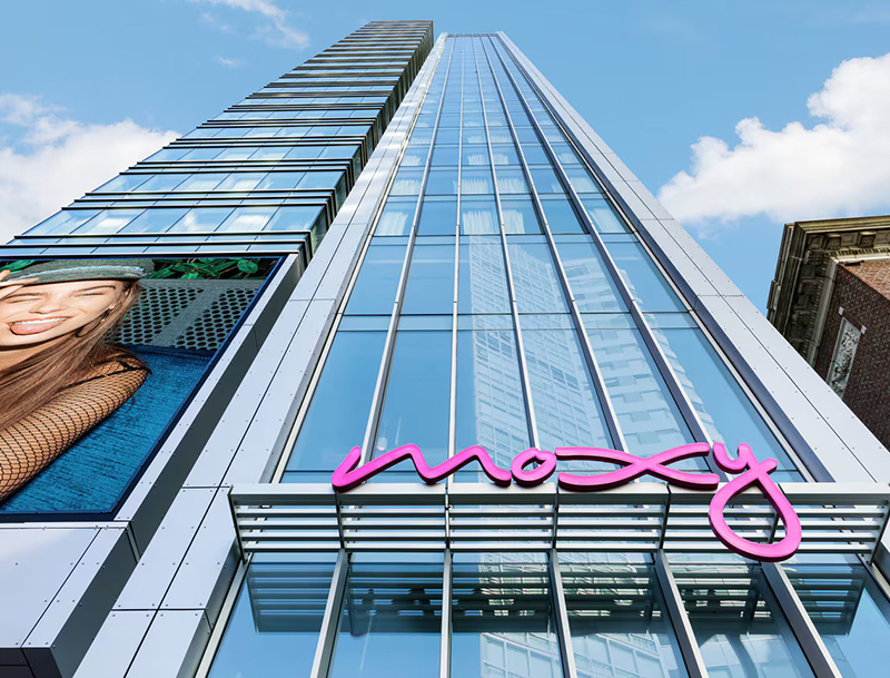 Moxy Boston Downtown's modern glass skyscraper exterior adorned with a vibrant pink neon sign and a large digital billboard of a woman.