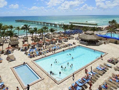 A birds-eye view of resort guests enjoying the pool and deck area, with a palm-dotted sandy beach and azure ocean waters visible in the distance.