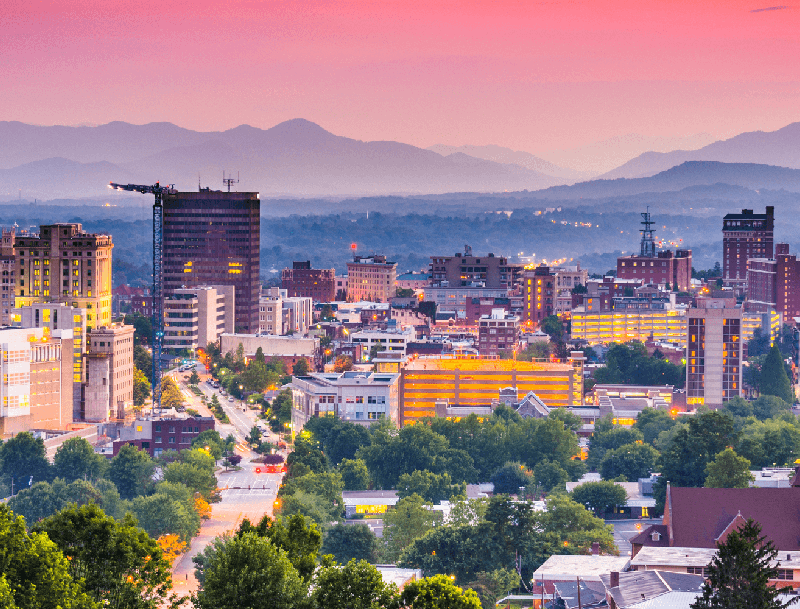 An elevated view of the Asheville, North Carolina skyline at dusk, with the Blue Ridge Mountains layered in the background under a pink and purple sky.