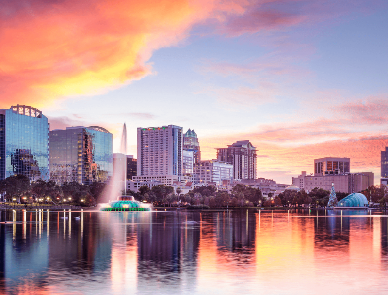 The city skyline of Orlando, Florida at sunset, featuring the illuminated Lake Eola fountain with vibrant orange and pink clouds reflecting on the calm water.