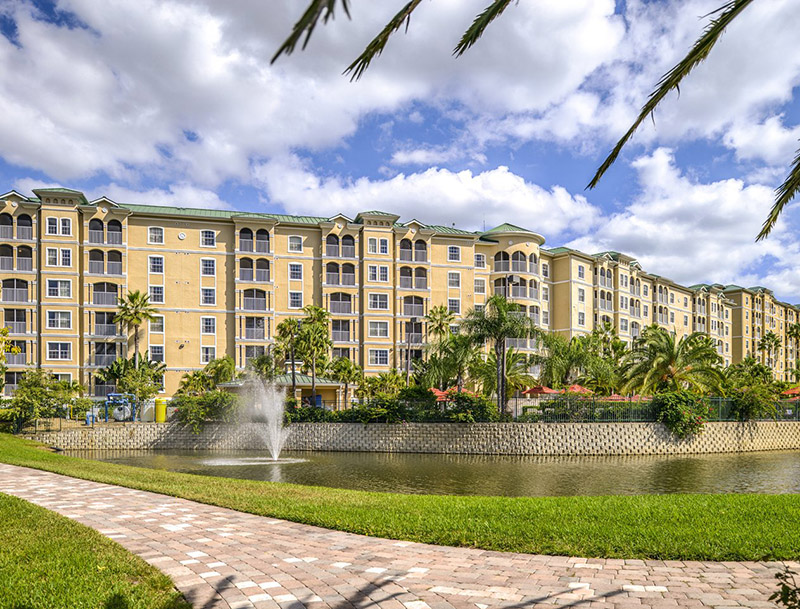 Multistory resort buildings with lush landscaping and a walking path along a lake in the foreground.