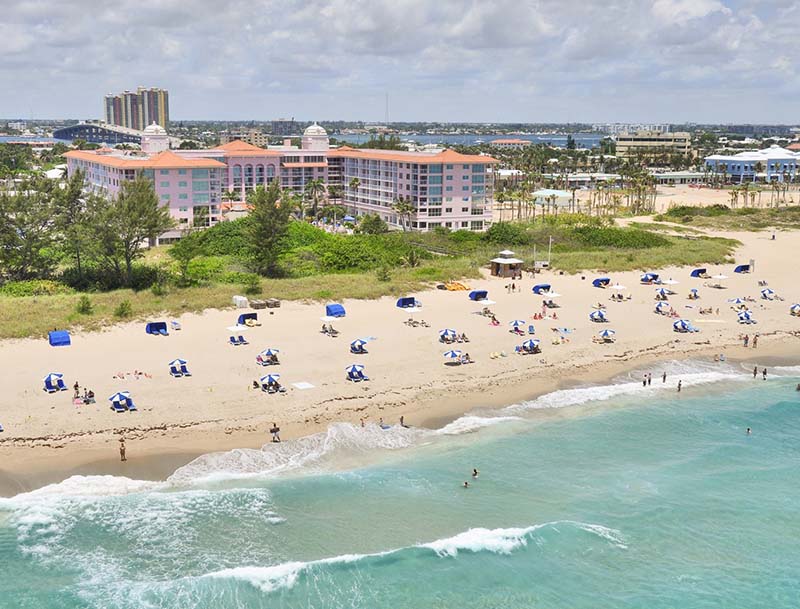 Aerial view of a sandy beach featuring sunbathers relaxing under colorful umbrellas, with a large resort visible in the background.