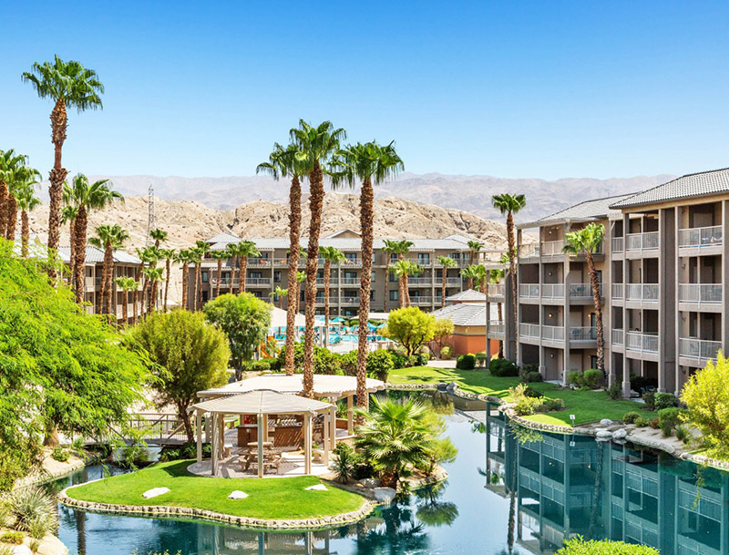 Multistory resort buildings surrounding a lushly-landscaped lake with mountains visible in the distance.