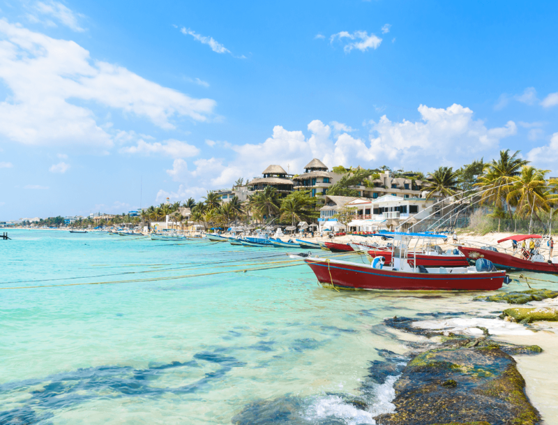 Several small fishing boats anchored in the clear turquoise waters along a sandy beach, lined with palm trees and tropical resorts.