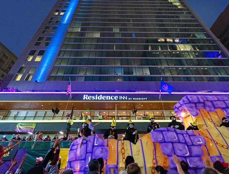 Low angle shot of the hotel's tall glass facade with a colorful Mardi Gras float passing in the foreground.