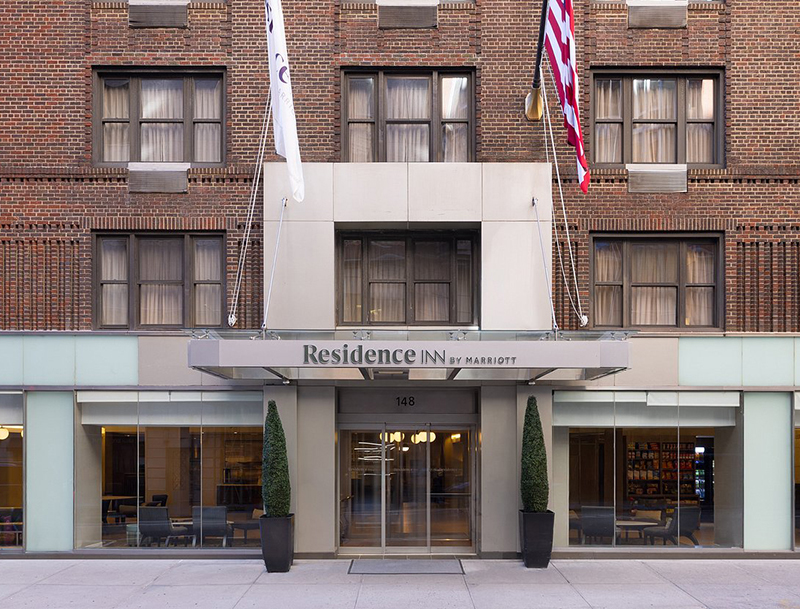 The hotel's street-level entrance, featuring a sleek glass and metal awning, tall potted topiaries, and large windows revealing the modern lobby.