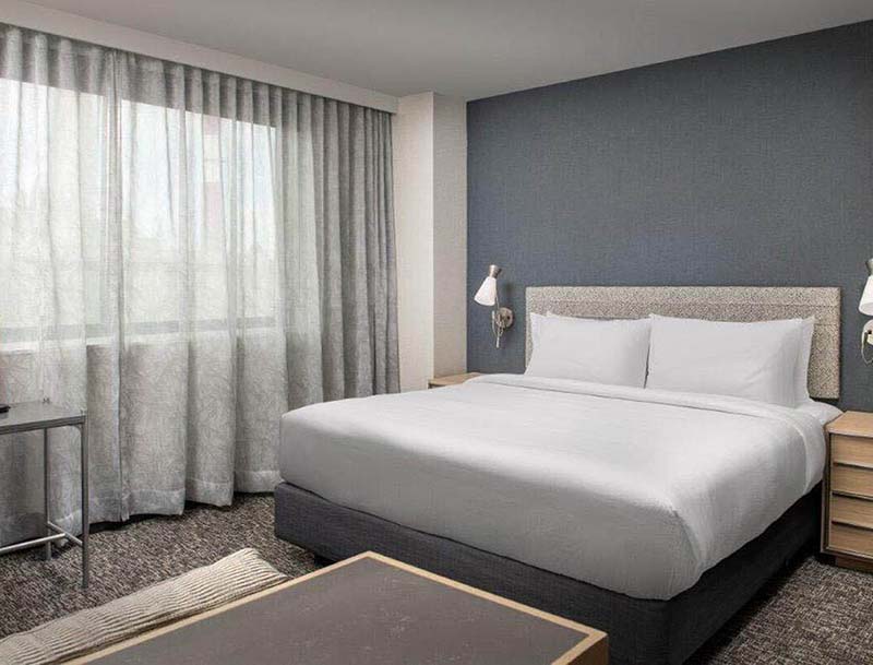 A serene hotel room in muted colors featuring a large bed, a desk and a seating area near a large curtained window.