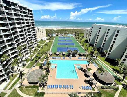 High-angle view of Royale Beach and Tennis Club, featuring a large swimming pool and blue tennis courts nestled between resort buildings leading to the beach.