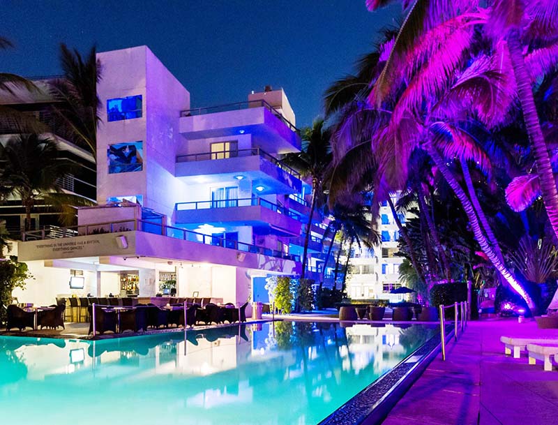 Night view of the hotel's illuminated facade and serene pool bathed in purple lights.
