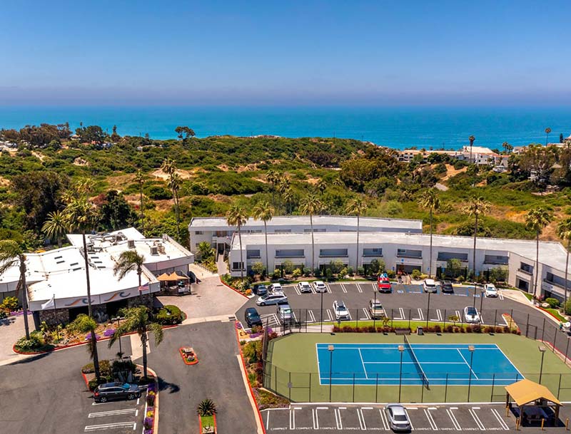 Aerial view of a tennis court surrounded by palm trees at a beachfront resort, with the ocean visible in the background.