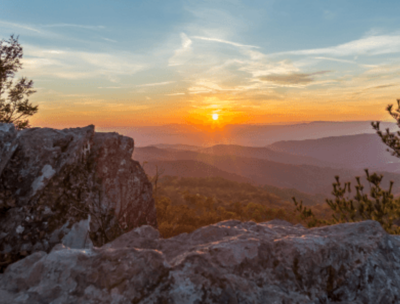 A scenic sunset view from a rocky overlook in Shenandoah Valley, Virginia, with the sun dipping below rolling mountain ridges.