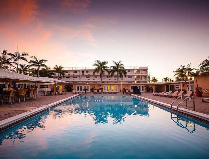 A large serene pool surrounded by lounge chairs and palm trees, with the resort building against the sunset sky in the background.