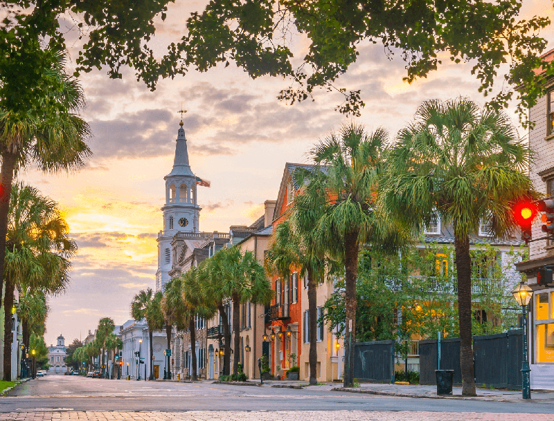 Historic Charleston street at sunset featuring palm trees, colorful row houses, and the St. Michael's Church spire.