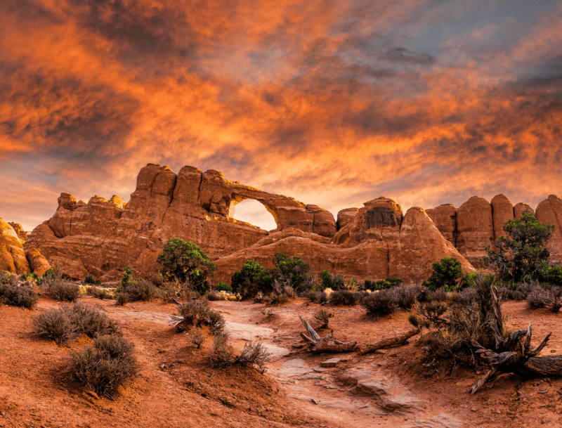 A natural red rock sandstone arch in Southern Utah under a dramatic orange and yellow sunset sky with desert brush in the foreground.