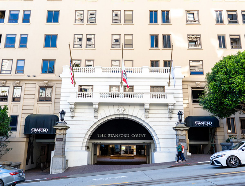Stanford Court's grand white arched entrance featuring decorative balconies, black awnings and flags as seen from the sloped city street.