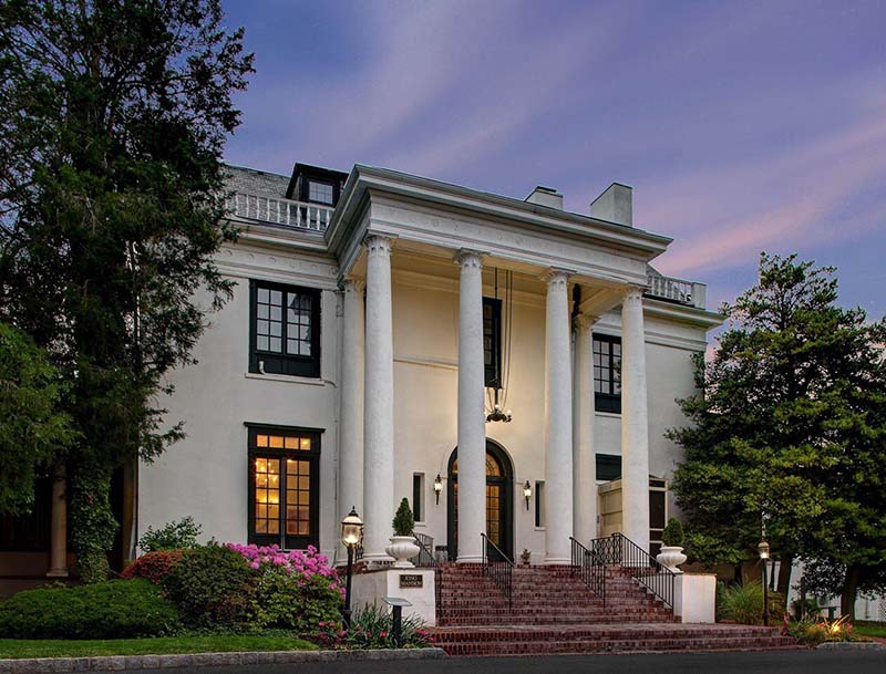 A view of Tarrytown House Estate's illuminated colonnade entrance and surrounding landscape at dusk.