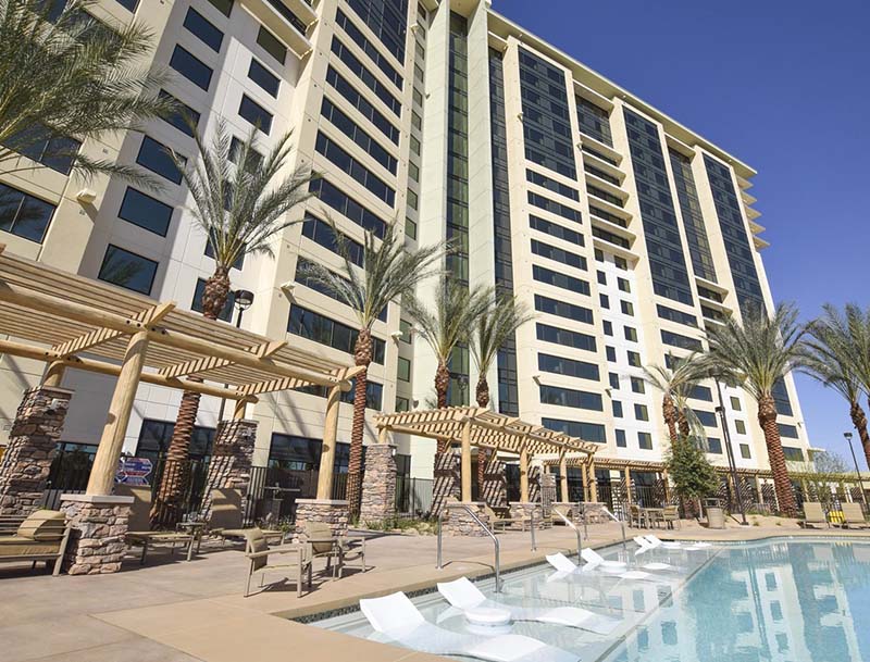 A low angle shot of the sunlit pool area featuring lounge chairs and tall palm  trees, with the resort building rising in the background.