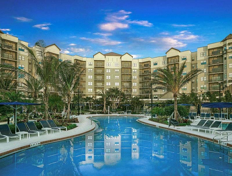 A large serene pool featuring lounge chairs and palm trees, with the multi-story resort buildings visible in the background.