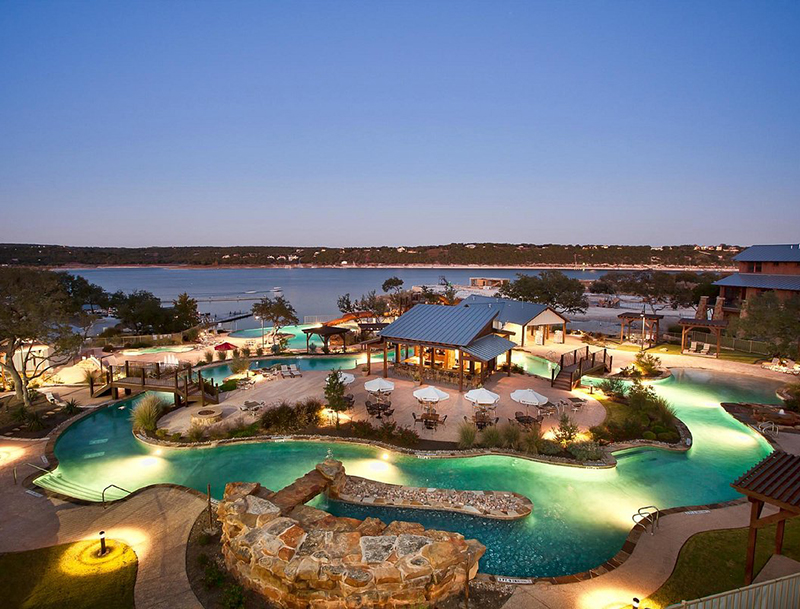 The resort's expansive pool area at dusk, featuring a winding illuminated lazy river, a poolside bar, and a scenic view of Lake Travis in the background.