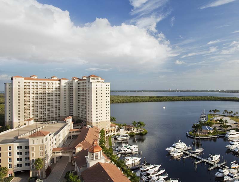 High-angle view of The Westin Cape Coral Resort at Marina Village, featuring a large hotel tower overlooking a marina filled with yachts and a vast nature preserve.