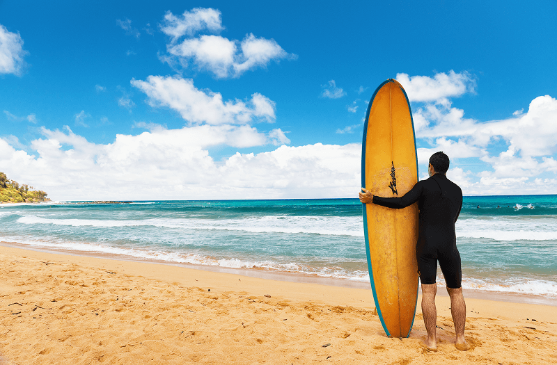 A man standing next to the ocean holding a brown surfboard on a bright day.