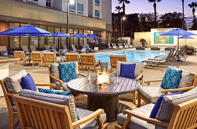 Poolside at the Cambria Hotel & Suites Anaheim with wooden chairs around a tabletop fire pit and a pool and lounge chairs in the distance.