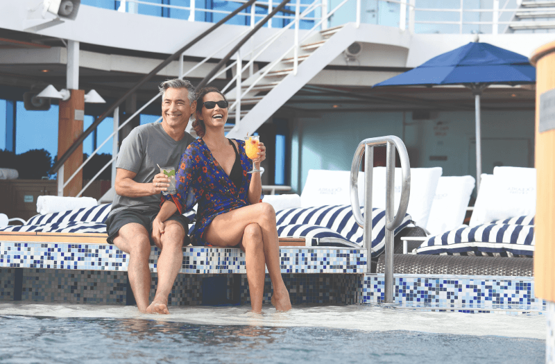 A couple enjoying drinks while they sit on the edge of a pool on an Oceania Cruises ship deck. 