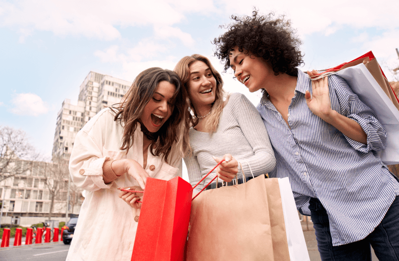 Three smiling friends looking into a gift bag while outside on a bright day.