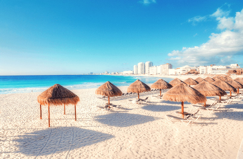A sunny beach scene featuring thatched umbrellas and lounge chairs, with hotels in the background.