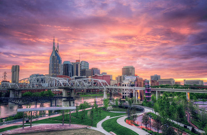 Nashville skyline at sunset with a view of a riverfront park and a bridge in the foreground.