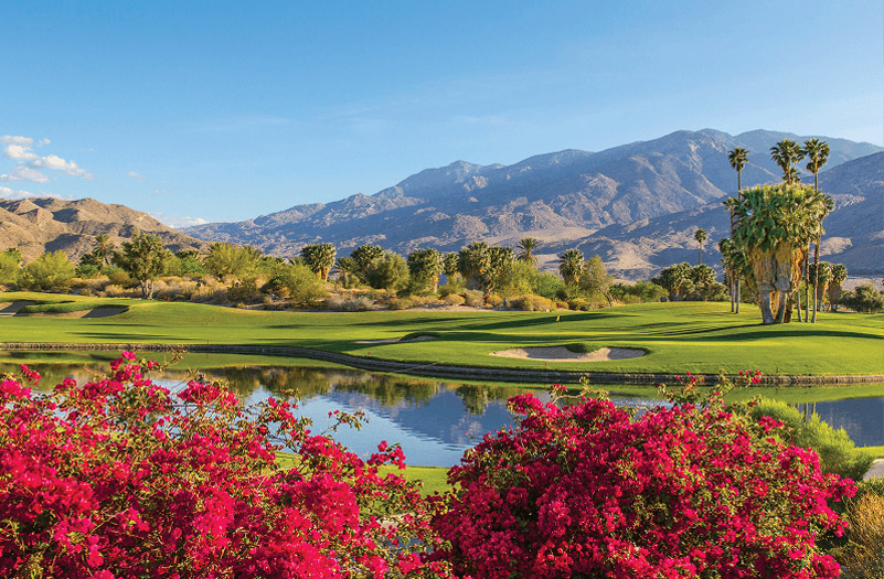A scenic golf course nestled among vibrant flowers in the foreground and majestic mountains in the distance.