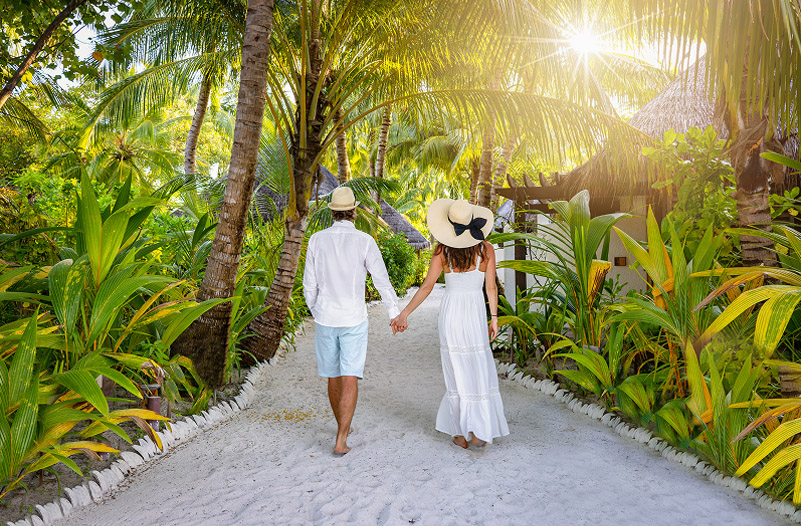 A man and woman walk hand in hand down a sandy path, surrounded by lush tropical greenery.