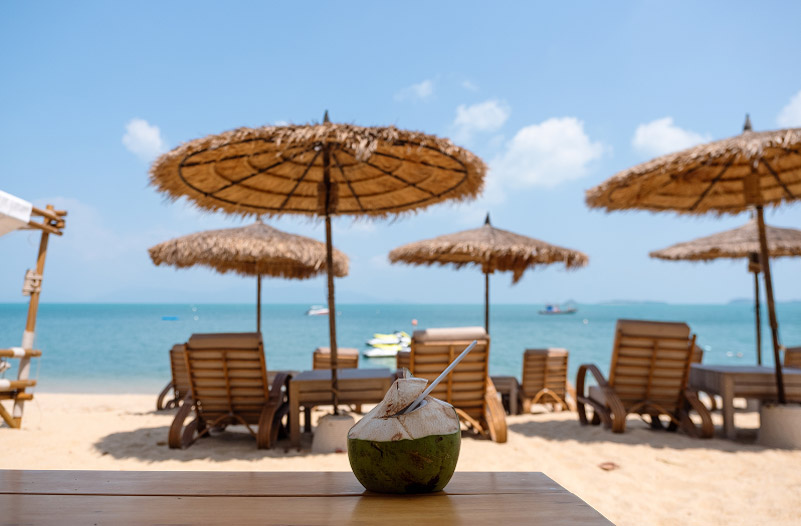 A coconut drink sits on a wooden table, with thatched umbrellas and lounge chairs lining the beach in the background.