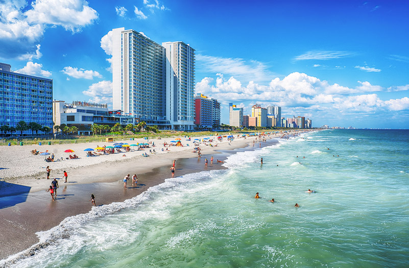 A lively beach scene with people swimming and sunbathing, and hotels lining the shoreline in the background.