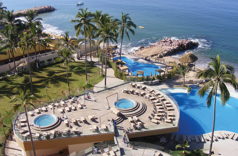 A serene resort-style pool surrounded by palm trees and a large deck, with a view of the ocean in the background.