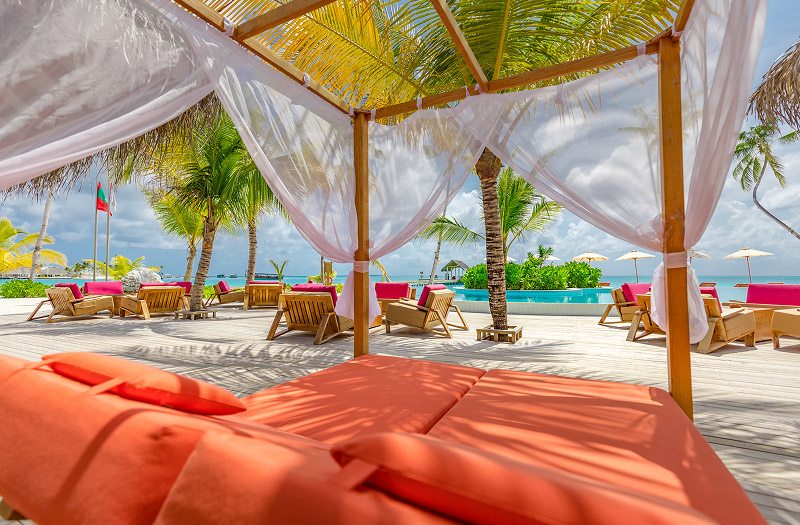 A vibrant orange pool bed on a shady resort-style pool deck with views of the pool, palm trees, and the ocean in the background.