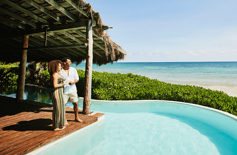 A couple standing on a wooden deck overlooking a private infinity pool, small green shrubs, and the ocean on a clear day.